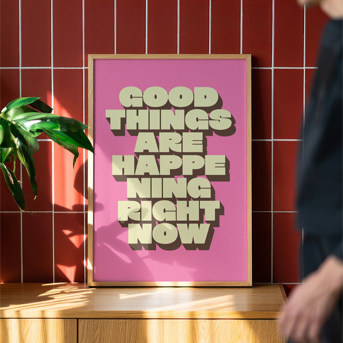 Framed poster with 'Good things are happening right now' text on a pink background, placed on a wooden surface against a red tiled wall.