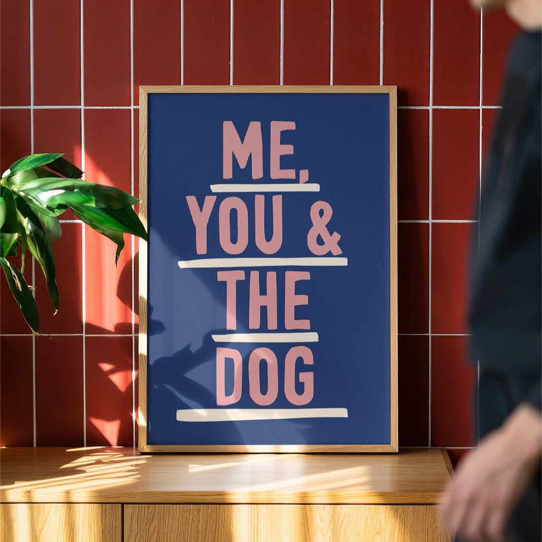 Framed poster with 'ME, YOU & THE DOG' text on a wooden surface with a red tiled wall background.