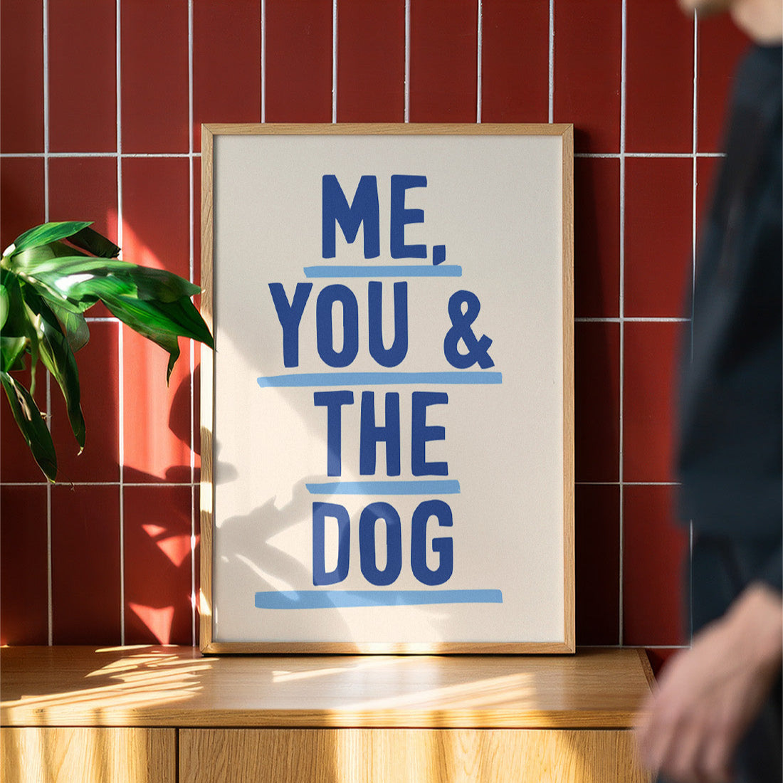 Framed poster with 'ME, YOU & THE DOG' text on a wooden surface with a red tiled wall background.