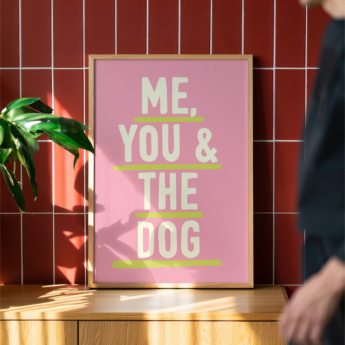 Framed poster with 'Me, You & The Dog' text on a pink background against a red tiled wall.