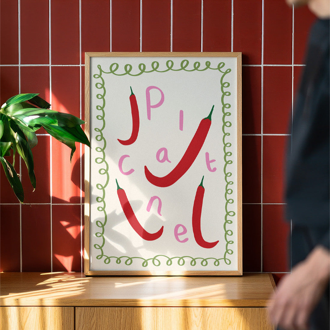 Framed artwork with red chili peppers and text on a wooden surface against a red tiled wall.