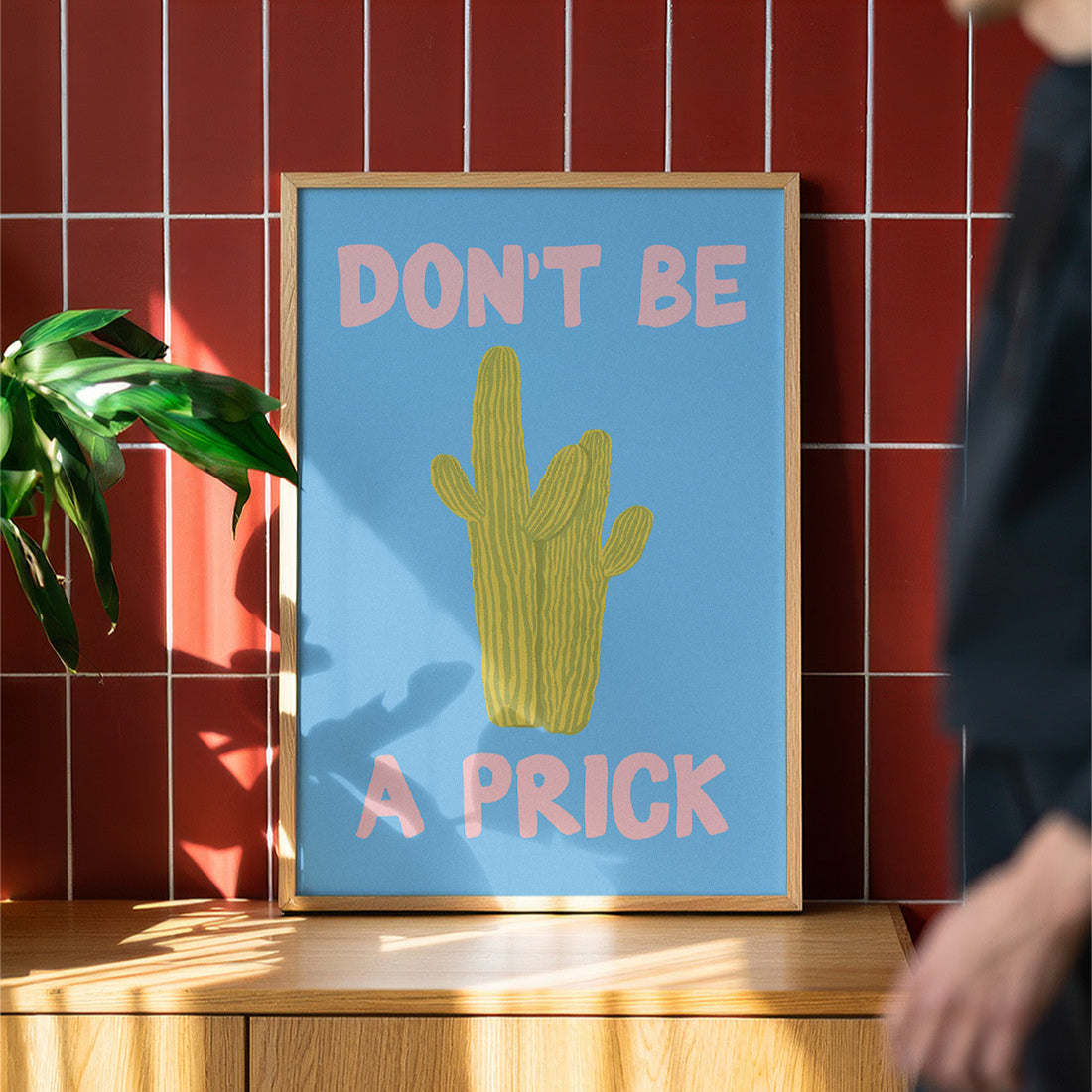 Framed poster with a cactus illustration and text on a wooden surface with a red tiled wall background.