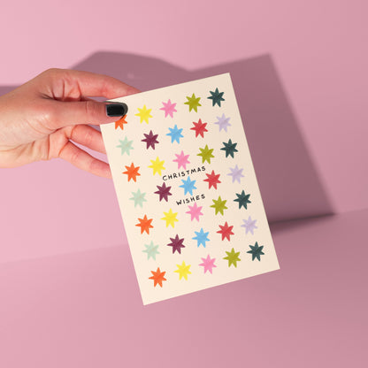 Hand holding a card with colorful star patterns and 'Christmas Wishes' text on a pink background