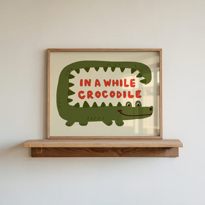 Framed artwork of a green crocodile with red text 'IN A WHILE CROCODILE' on a wooden shelf.