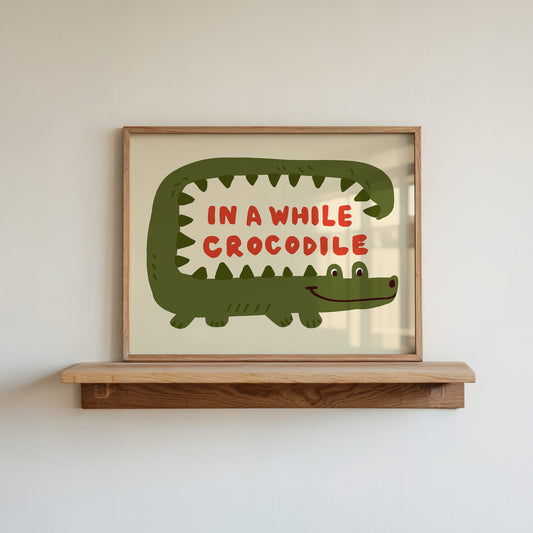Framed artwork of a green crocodile with red text 'IN A WHILE CROCODILE' on a wooden shelf.