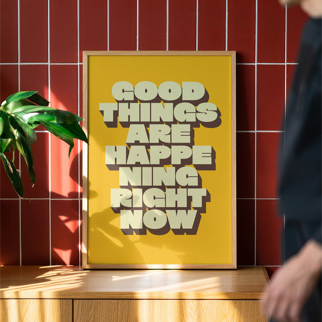Yellow framed poster with 'Good things are happening right now' text on a wooden surface with a red tiled wall background.