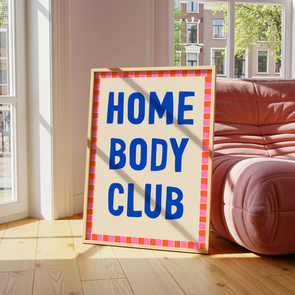 Framed sign with 'HOME BODY CLUB' text in a room with a red sofa and large windows.