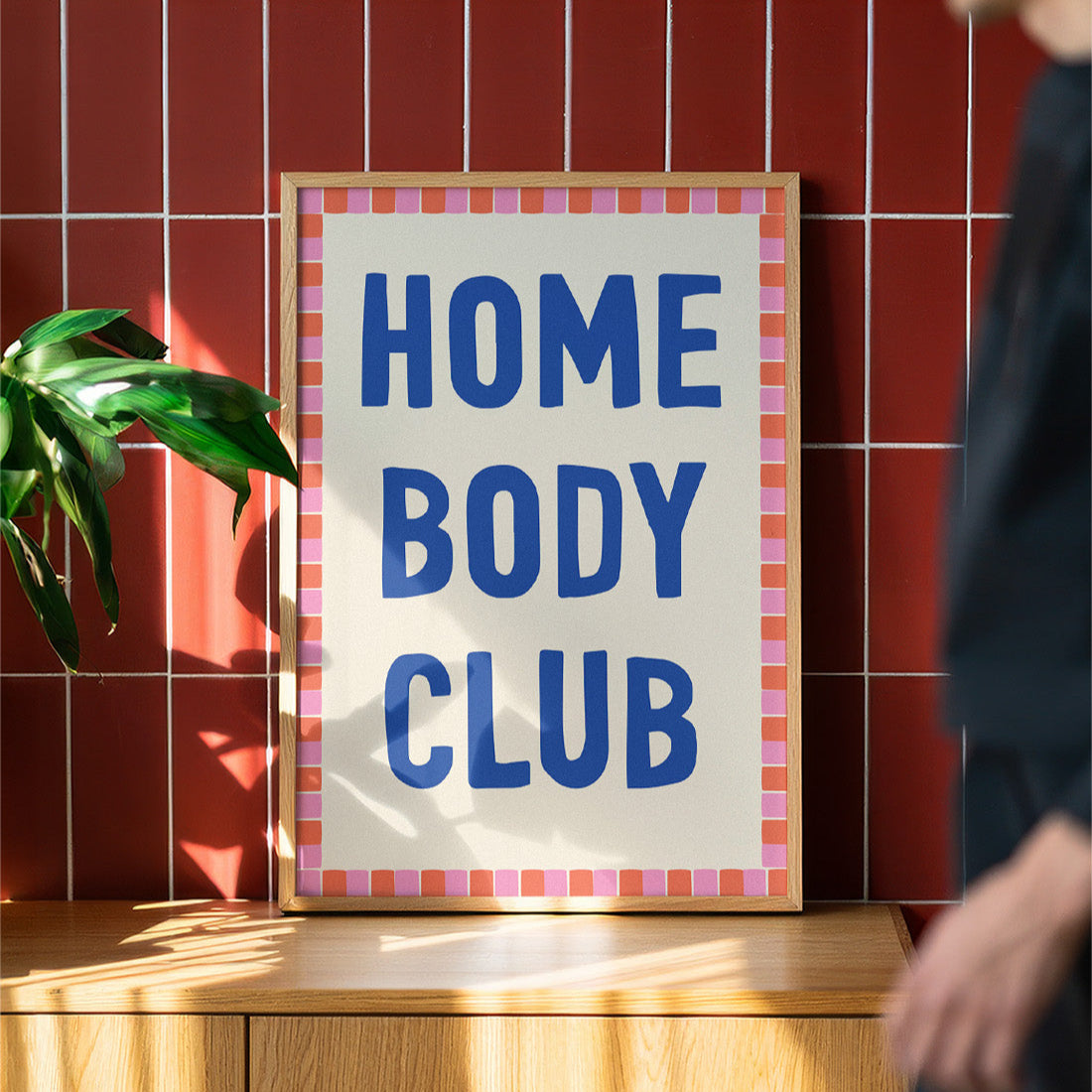 Framed sign with 'HOME BODY CLUB' text on a wooden surface against a red tiled wall.